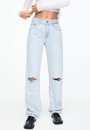 Light blue distressed jeans with ripped knees, featuring a high-rise waist and wide-leg fit. Paired with black studded slip-on shoes.