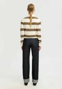 Žena s plavom repom koja nosi dugi pamučni majicu u krem i smešnim prugama, tamne široke hlače s manžetama i smešnim cipelama, okrenuta leđima.