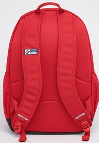 Rode rugzak met gevoerde schouderbanden, nylon textuur, logo embleem, zijzakken en een zwarte onderkant, met een compact en gestructureerd ontwerp.