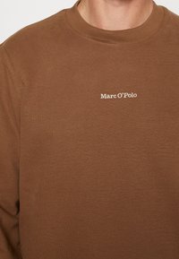Sweat-shirt marron à encolure ras-du-cou avec le logo blanc « Marc O'Polo » brodé sur le côté gauche de la poitrine.