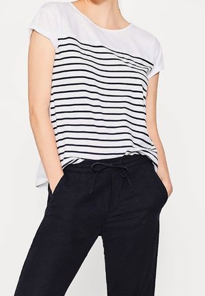 Personne portant une chemise blanche à manches courtes avec des rayures horizontales noires et un pantalon noir avec cordon de serrage, les mains dans les poches.