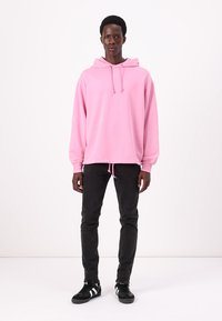 Hoodie oversized rose avec des manches longues et un cordon de serrage, associé à un jean noir slim et des baskets noires avec des accents blancs.