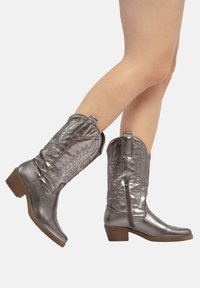 Botas de vaquero metálicas plateadas con tacón cuadrado, costuras decorativas, lengüetas para tirar y un detalle de cremallera lateral. Textura de cuero suave.