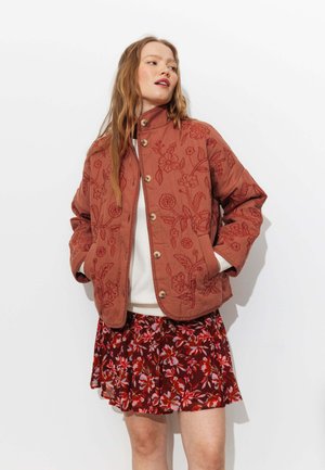 Femme portant une veste matelassée couleur rouille avec broderies florales, un haut blanc et une jupe plissée à fleurs rouges, debout devant un fond blanc.