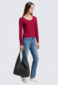 Langarm geribd top in karmozijnrood met knoopdetail, gecombineerd met lichtblauwe skinny jeans en een grote zwarte hobo-tas. Witte sneakers maken de look compleet.