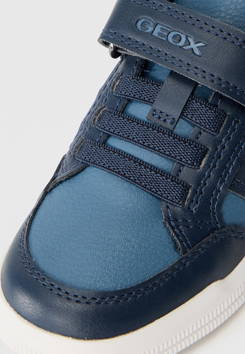 Baskets en cuir bleu foncé avec un design texturé. Dotées de lacets élastiques, d'une sangle Velcro et d'une semelle en caoutchouc blanche. Marque GEox visible.