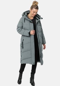 Abrigo acolchado largo y gris con capucha, que presenta bolsillos con cremallera y textura acolchada. Llevado sobre un conjunto negro y botas negras hasta la rodilla.