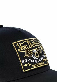 Schwarze Baseballmütze mit einem bestickten goldenen und weißen Patch, der ein Auge mit Flügeln, Text und dekorativen Blitzmotiven zeigt.