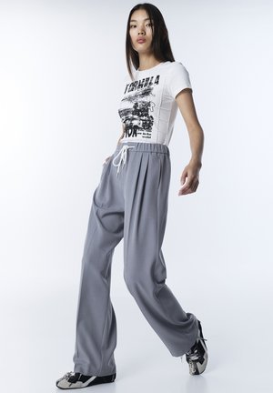 White T-shirt with black print, paired with light grey pleated trousers and black sneakers featuring decorative hardware details.