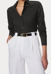 Camisa negra de botón y mangas largas combinada con pantalones blancos de tiro alto, sujetos por un cinturón negro con una hebilla ovalada dorada.