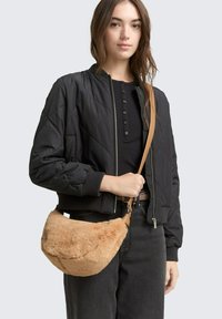 Schwarze, gesteppte Bomberjacke mit gerippten Bündchen, kombiniert mit einer braunen, gefälschten Fell-Umhängetasche mit Schulterriemen. Dunkle Jeans runden den Look ab.