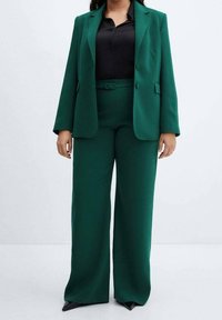 Abito verde su misura con blazer strutturato a due bottoni e pantaloni a gamba larga. Indossato sopra una camicia nera, presenta una texture liscia.