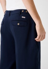 Marineblaue Hose mit zwei Gesäßtaschen, braunen Knöpfen und einem kleinen Markenetikett. Glatte Textur mit maßgeschneiderter Passform.