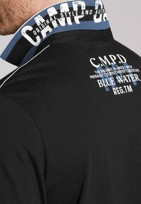 Czarna koszulka polo z niebieskim i białym prążkowanym kołnierzykiem. Posiada nadrukowany tekst na kołnierzyku i z tyłu, w różnych rozmiarach i kolorach.