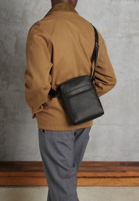 Borsa a tracolla nera in pelle con finitura texturizzata, forma rettangolare e tracolla regolabile, indossata sopra una giacca marrone e pantaloni grigi.
