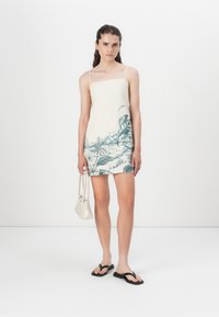 Linnen mini-jurk met dunne bandjes, voorzien van een groen tropisch landschap print op een crèmekleurige achtergrond. Draagt met zwarte teenslippers en een witte tas.