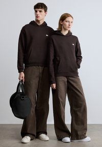 Καφέ φούτερ με κουκούλα και μπροστινές τσέπες, συνδυασμένα με καφέ φαρδιά παντελόνια. Μια μαύρη στρογγυλή τσάντα χειρός με κοντά χερούλια κρατιέται από μια φιγούρα.