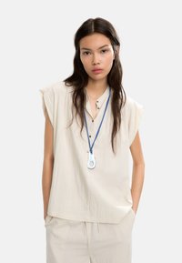 Beżowa, luźna bluzka z krótkimi rękawami, z zapięciem na guziki, połączona z pasującymi spodniami. Posiada niebieską smycz z charakterystycznym wisiorkiem.