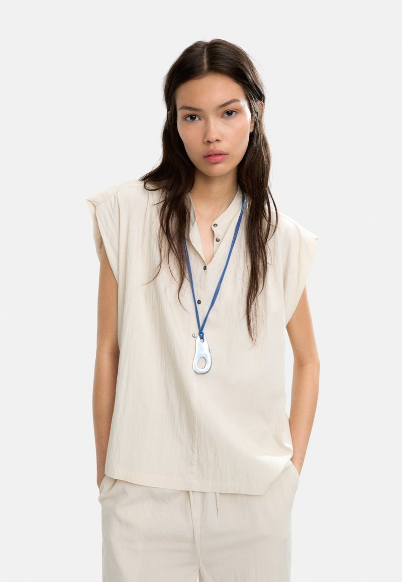 Beżowa, luźna bluzka z krótkimi rękawami, z zapięciem na guziki, połączona z pasującymi spodniami. Posiada niebieską smycz z charakterystycznym wisiorkiem.