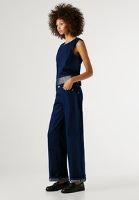 Tmavomodrý denimový overal so širokými nohavicami, bez rukávov, striebornými akcentmi v oblasti pásu a lemami na nohách. Lesklé čierne poltopánky doládzajú celkový vzhľad.