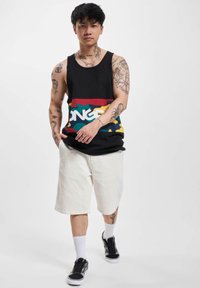 Svart tanktop med flerfarget camouflasjemønster og hvit skrift, kombinert med hvite kordfløyelsshorts og svarte sneakers med hvite såler.