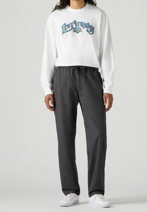 Osoba nosząca biały sweter Levi Strauss, luźne ciemnoszare spodnie i białe trampki stoi na jednolitym tle.