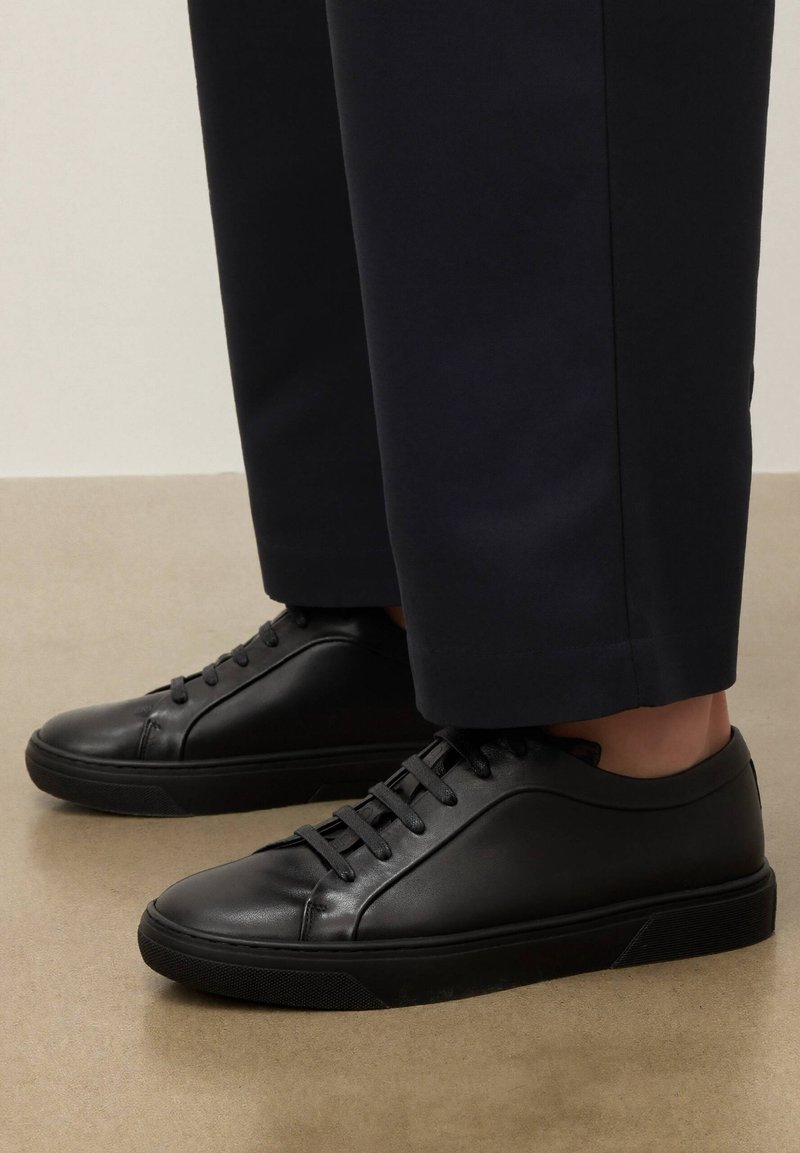 Adidași negri din piele, cu talpă joasă și șireturi negre, purtați cu pantaloni negri scurtați, pe un podea bej.