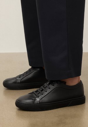 Mustad madala säärega nahast tossud mustade paeltega, kantud koos lühendatud mustade pükstega beežil põrandal.