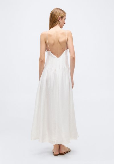Žena s rovnými blond vlasmi, oblečená v dlhých bielych šatách bez rukávov s tenkými ramienkami a otvoreným hlbokým chrbtom, stojaca na bielom pozadí.