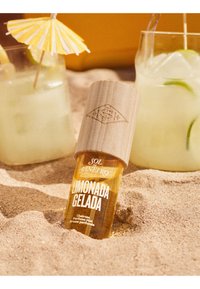 Botella de perfume en spray Limonada Gelada parcialmente enterrada en la arena junto a dos bebidas heladas de limón-lima, una de ellas con un paraguas de papel amarillo.