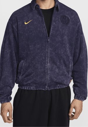 Mand iført en mørkeblå jakke med lynlås med et gult Nike-logo og broderet cirkulært emblem på øverste del af brystet.