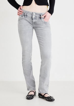 Personne portant un jean gris délavé, les mains dans les poches et des ballerines noires à clous sur fond blanc.
