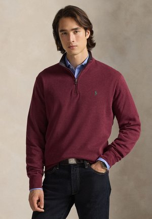 Jeune homme aux cheveux foncés portant un pull bordeaux à fermeture éclair quart de zip sur une chemise bleue à col et un jean foncé, une main dans la poche.