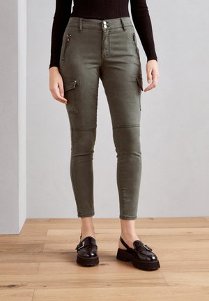 Zelené cargo kalhoty ve slim fit střihu, s kapsami na zip a panely na kolenou, kombinované s černými botami na silném podpatku.