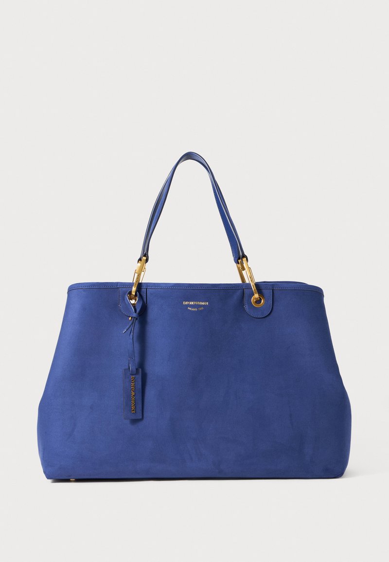 Sac fourre-tout en suédine bleue avec quincaillerie en métal doré, doubles poignées supérieures et une étiquette de marque. Présente une forme structurée et une texture lisse.