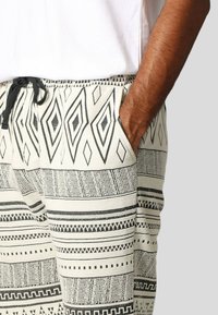 Katoenen broek met een crème basis en zwarte geometrische patronen, waaronder diamanten en strepen, en een trekkoord in de taille voor verstelbaarheid.