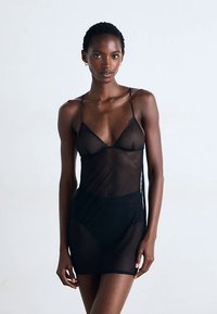 Fekete áttetsző fehérnemű vékony pántokkal, testhezálló kialakítással és félig átlátszó anyaggal, kiemelve a sziluettet.