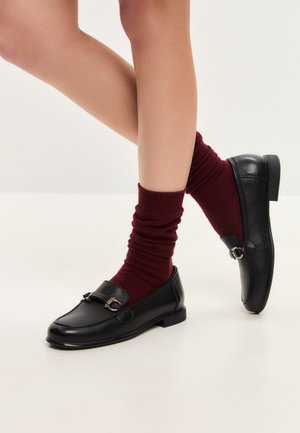 Black leather loafers with a round toe, accentuated by a metal buckle detail, paired with burgundy slouchy socks.