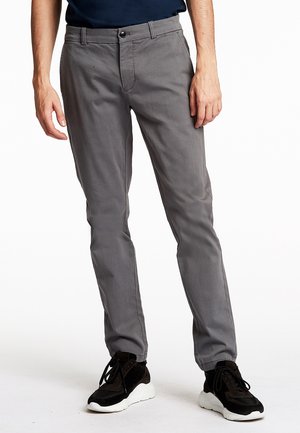 Mann trägt eng anliegende graue Hose und schwarze Sneakers mit weißen Sohlen, steht vor weißem Hintergrund.