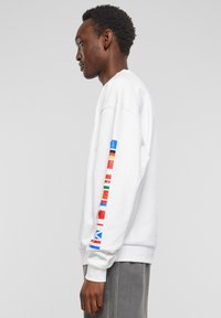 Hvit sweatshirt med lange ermer som har fargerike flaggdesign langs armen. Laget av myk stoff med en avslappet passform og ribbet mansjetter.