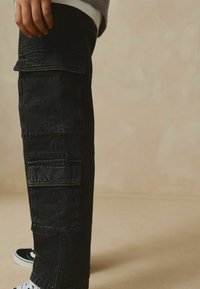 Pantalons cargo en denim noir avec plusieurs poches latérales, une coupe décontractée et des détails de couture contrastants, associés à des baskets blanches.