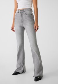 Jean gris évasé en denim, avec une taille haute, des poches avant et des ourlets fendus. Associé à des chaussures noires pointues.