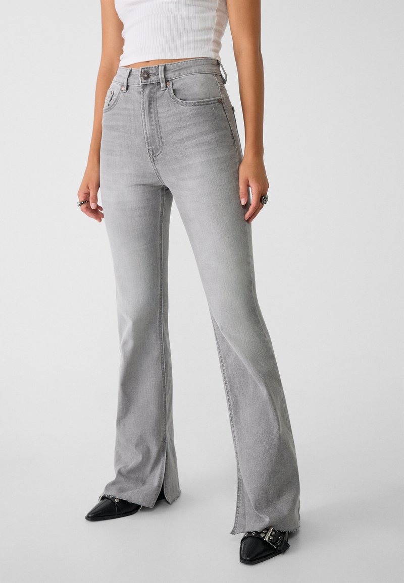 Jean gris évasé en denim, avec une taille haute, des poches avant et des ourlets fendus. Associé à des chaussures noires pointues.