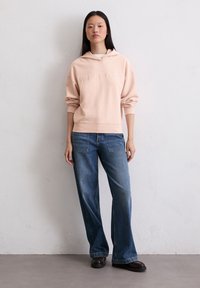 Sweatshirt rosa chiaro con cappuccio e logo in rilievo, abbinato a jeans blu oversize e scarpe marrone scuro; design semplice e informale.
