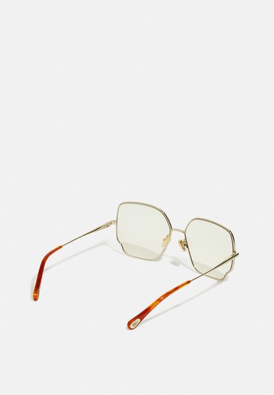 Ochelari de soare cu ramă aurie, cu lentile hexagonale, având lentile de culoare verde deschis și un model de țestoasă pe vârfurile brațelor.