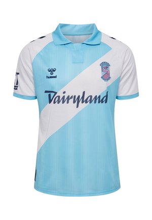 Ung person iført en lyseblå og hvid sports-trøje, denimshorts, hvide sokker og sorte sneakers med pink detaljer, stående mod en ensfarvet baggrund.