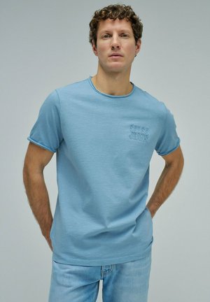 Hombre con cabello rizado que lleva una camiseta azul claro de "Salsa Jeans" y vaqueros azul claro, de pie con las manos detrás de la espalda, fondo neutro.