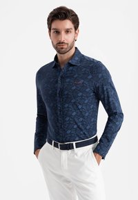 Navy bloemenoverhemd met lange mouwen, een button-down kraag en subtiele rode borduursels. Gecombineerd met witte broeken en een donkere riem.