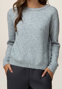 Pull en maille gris clair avec de petits clous argentés le long de l'encolure et des manches, porté avec un pantalon foncé, mains dans les poches.