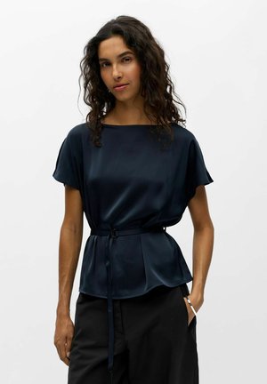 Femme aux cheveux bouclés portant un chemisier court en satin bleu marine avec une ceinture à la taille et un pantalon noir, debout avec une main dans la poche.